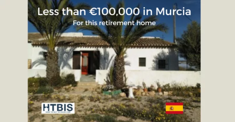 A retirement home in Murcia priced at less than €100,000. The image shows a white house with a tiled roof, palm trees in the front yard, and a Spanish flag at the bottom right corner. This Murcia property investment is perfect for those looking to retire amidst the serene beauty of Mazarron real estate.