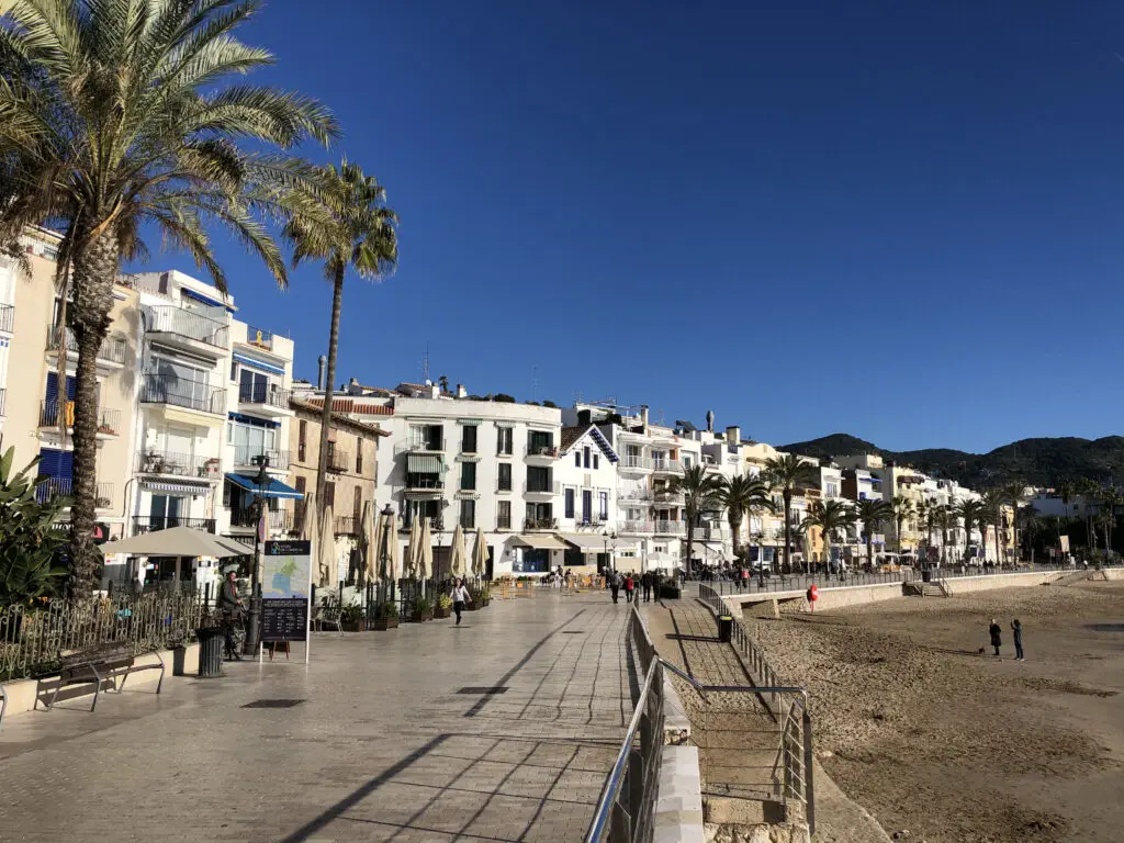 Seaside promenade with palm trees, outdoor cafes, and colorful buildings beside a sandy beach under a clear blue sky—a perfect spot to enjoy the best things to do on the Costa de Barcelona.