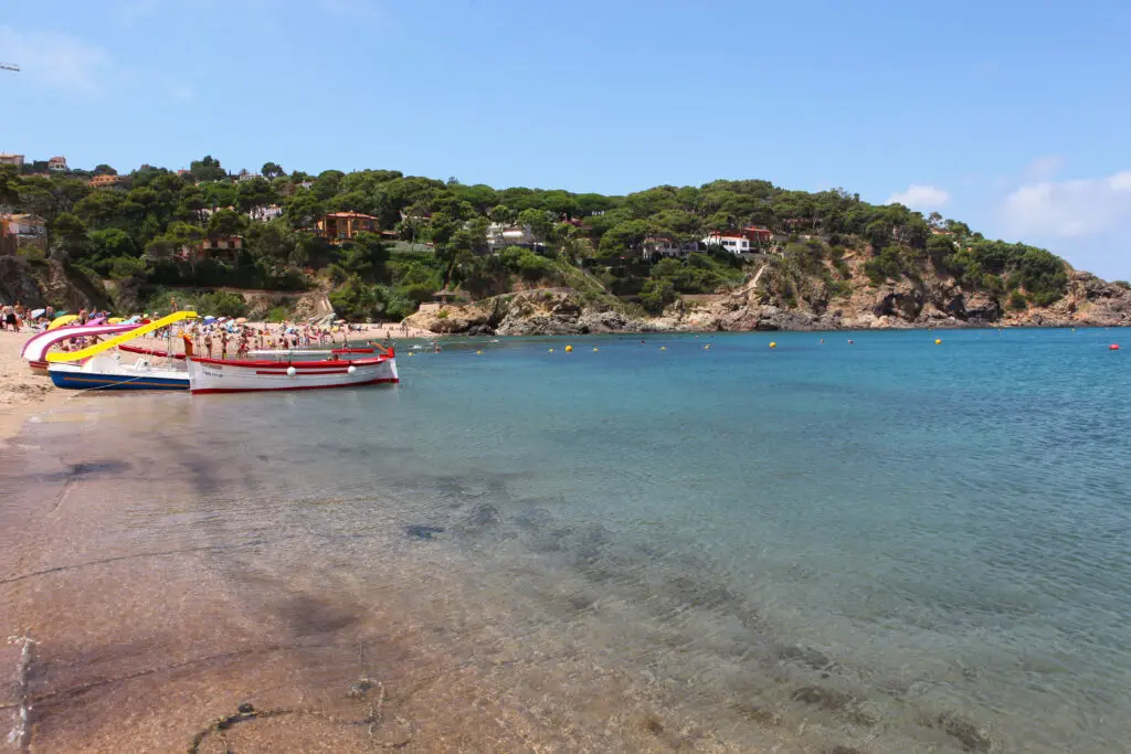 Several small boats are docked along a sandy beach in Costa Brava Spain, with people sunbathing nearby and green hills with houses in the background beside a clear, calm sea.