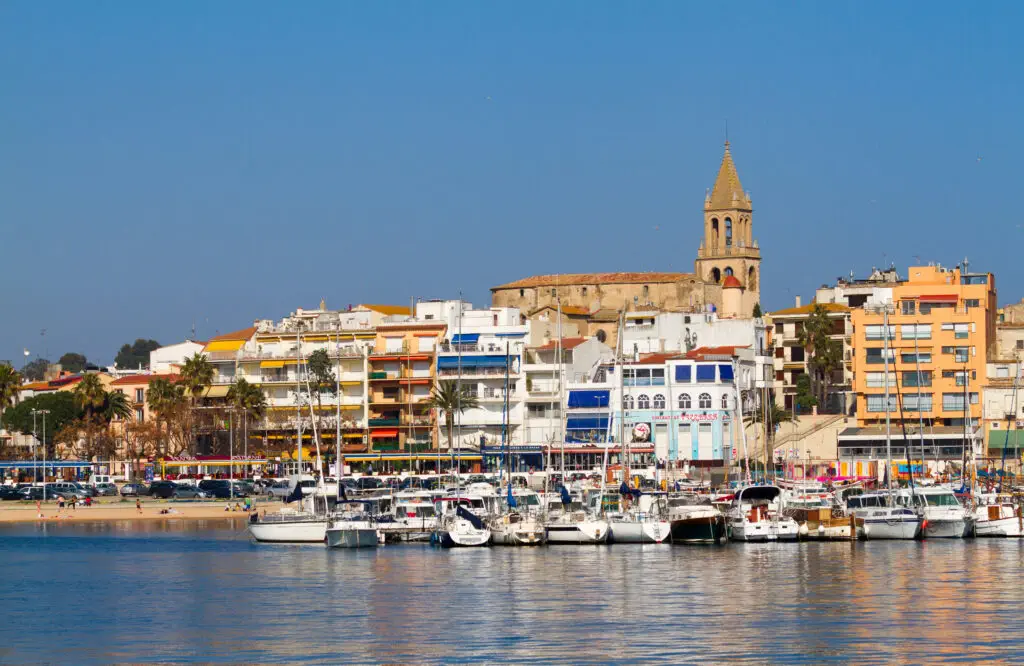 A waterfront view of colorful buildings and a church tower behind a marina filled with docked boats under a clear blue sky in Costa Brava, Spain.