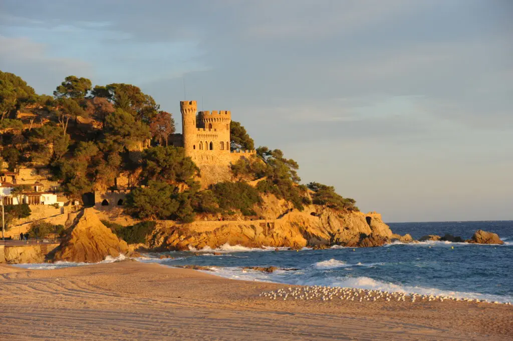 A stone castle-like building stands on a rocky, tree-covered hill overlooking a sandy beach and the ocean under a clear sky along the stunning Costa Brava Spain.