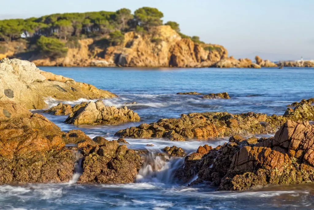 Rocky coastline with ocean waves gently flowing over the rocks; an island with trees is visible in the background under a clear sky, capturing the scenic beauty of Costa Brava in Spain.