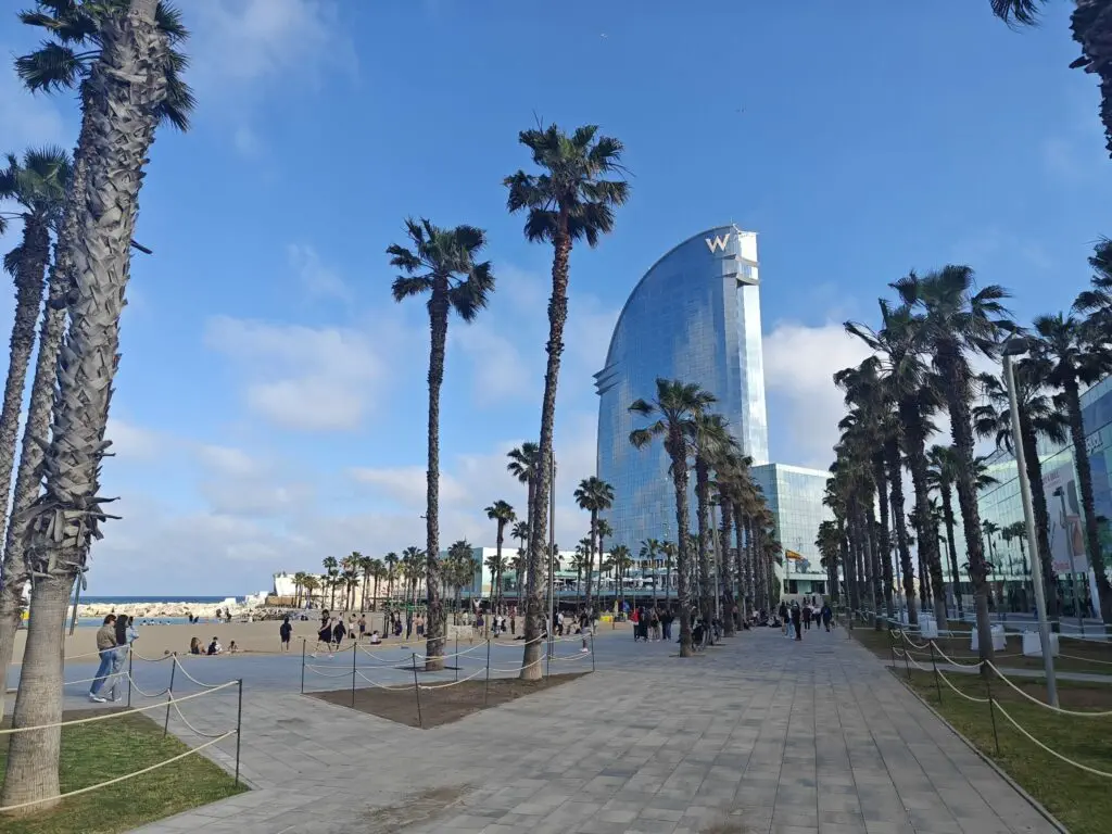 Palm trees line a walkway near a sandy beach bustling with people; a tall, sail-shaped glass building rises in the background—an iconic spot often featured in buying property in Spain guides and discussions on holiday rental Spain regulations.