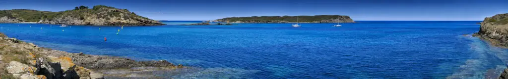 Panoramic view of a calm blue sea with a rocky coastline, small islands, and anchored sailboats under a clear sky—one of the best things to do in Balearic Islands for nature lovers and sailing enthusiasts.