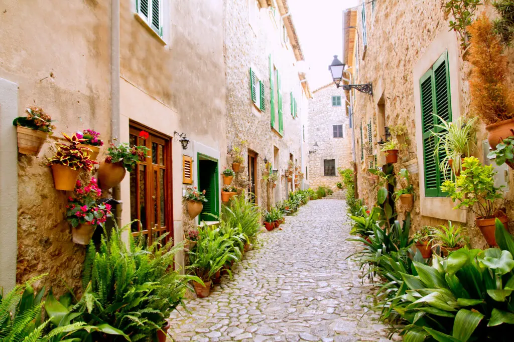 A narrow cobblestone street lined with stone buildings, green shutters, and numerous potted plants evokes the timeless charm found in many Balearic Islands villages.