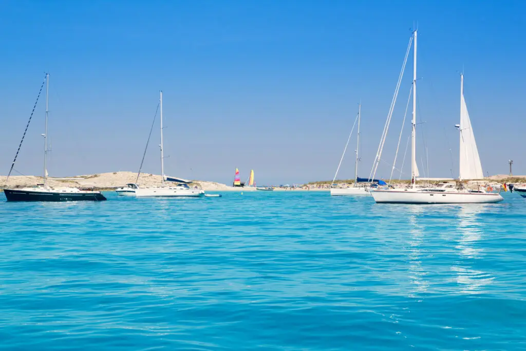 Several sailboats float on calm, clear blue water near a sandy shoreline under a bright blue sky, evoking the tranquil beauty often found in the Balearic Islands.
