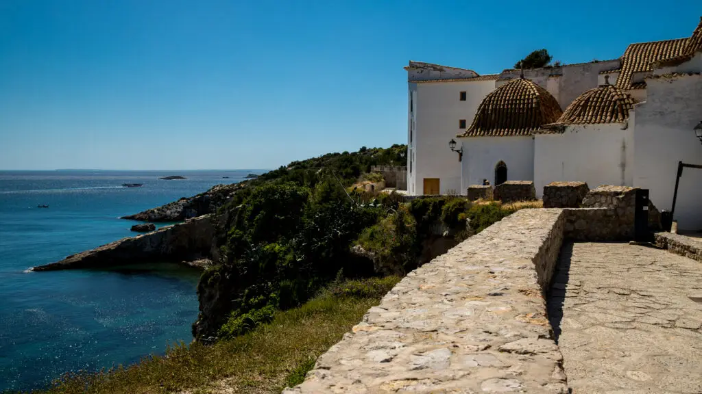 A stone path winds along a coastal cliff beside a white building with domed roofs, overlooking the blue sea—a scene straight from the Balearic Islands map and one of the best things to do in the Balearic Islands.