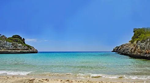 A sandy beach with blue water and a rocky cliff.
