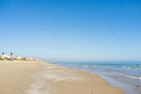 A wide sandy beach with gentle waves under a clear blue sky stretches along the Costa Blanca. Few buildings are visible in the distance.
